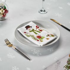 a white plate topped with a snowman napkin next to a fork and glass filled with christmas decorations