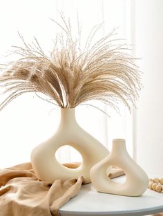 two white vases sitting on top of a table with dried grass in the middle