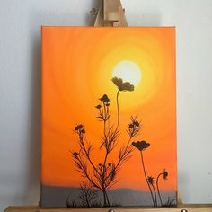 a painting on a easel with the sun setting in the background and flowers growing out of it