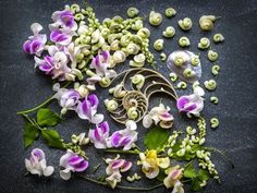 purple and white orchids are arranged on a black surface next to green leaves, beads and other flowers