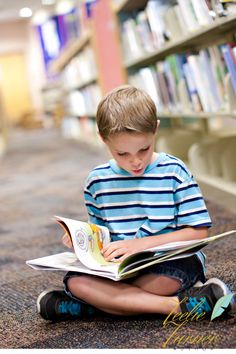 a young boy sitting on the floor reading a book in a library with bookshelves behind him