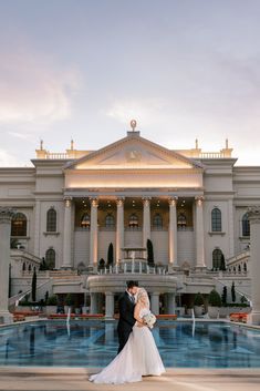 a bride and groom standing in front of a large building with a fountain at sunset