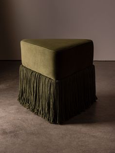 a green footstool with fringes on the top and bottom, in front of a gray wall