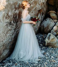 a woman in a wedding dress standing next to a large rock and holding a bouquet