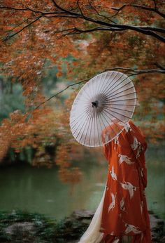 a woman in an orange and white kimono is standing by the water with her umbrella up