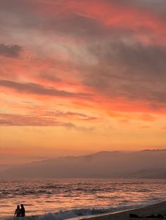 two people are sitting on the beach watching the sun set over the ocean and mountains