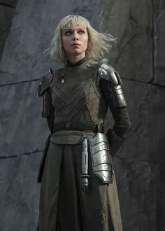 a woman with blonde hair and armor standing in front of a stone wall, looking at the camera