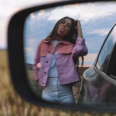 a woman standing in front of a car looking at her reflection in the rear view mirror