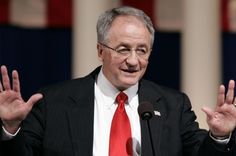 a man in a suit and red tie giving a speech with his hands raised up