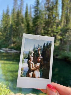 a person holding up a polaroid photo in front of a lake with trees on the other side