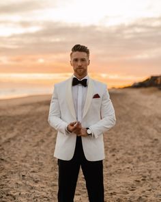 a man in a white tuxedo standing on the beach at sunset with his hands in his pockets