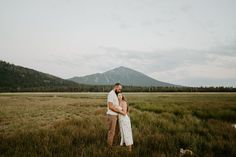 a man and woman standing in the middle of a field with mountains in the background