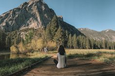 a woman sitting on a wooden bridge in front of a mountain with trees and grass