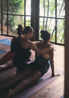 two women are sitting on yoga mats in a room with wooden floors and large windows