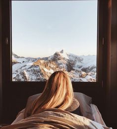 a woman sitting in bed looking out the window at snow covered mountains and hills outside