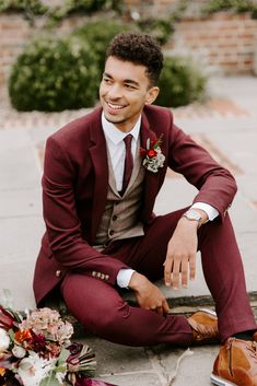 a man in a suit and tie sitting on the ground next to some flowers, smiling