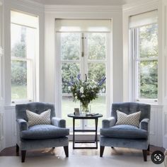 two chairs and a table in front of three large windows with flowers on the table