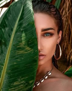 a woman with blue eyes and large hoop earrings is posing behind a green palm leaf