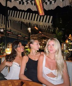 three beautiful women sitting at a table in front of a bar with lights hanging from the ceiling