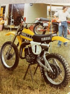 a yellow dirt bike parked on top of a grass covered field next to a man