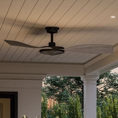 an outdoor ceiling fan mounted on the side of a house's porch with trees in the background