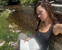 a woman sitting on a rock next to a river and holding a book in her hand