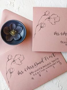 two pink envelopes with black ink on them and a flower in a blue bowl