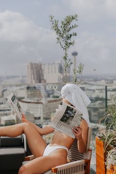 a woman sitting on top of a wooden chair next to a plant and reading a newspaper