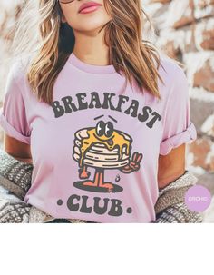 Embrace the joy of mornings with our 'Breakfast Club' tee, where comfort meets whimsy! Crafted from soft, cozy fabric, this shirt features an adorable stack of pancakes, complete with a buttery grin that'll brighten your day. Perfect for lazy brunches or just lounging around, it's a deliciously delightful addition to your wardrobe. Join the club and spread the pancake happiness everywhere you go! Graphic Tee With Letter Print For Brunch, Funny Print Graphic Tee For Brunch, Screen Print Crew Neck T-shirt For Brunch, Crew Neck T-shirt With Screen Print For Brunch, Funny Print Short Sleeve T-shirt For Brunch, Funny Print Cotton Tops For Brunch, Cute Crew Neck Tops For Brunch, Cotton Top With Funny Print For Brunch, Graphic Tee With Screen Print For Brunch