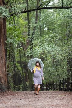 a pregnant woman walking in the woods with an umbrella over her head and holding a yellow scarf around her neck