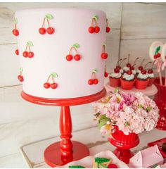 a white cake with cherries on it sitting next to cupcakes and flowers