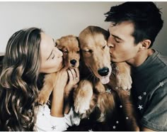 a man and woman kissing while holding two puppies in their lap, with the dog's tongue hanging out