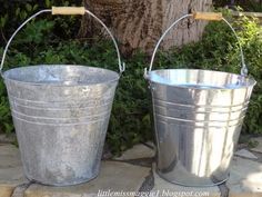 two metal buckets sitting next to each other on a stone floor near a tree