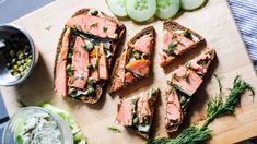 a wooden cutting board topped with slices of bread covered in salmon and cucumbers