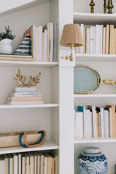 a white book shelf filled with books next to a lamp and vase on top of it