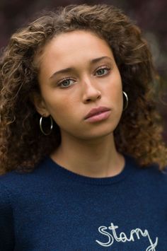 a woman with curly hair wearing a blue shirt and hoop earrings is looking at the camera