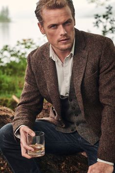 a man in a brown jacket and tie holding a drink sitting on a tree stump