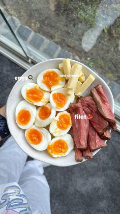 a person holding a white plate with eggs, meat and cheese on it in front of a window