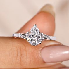 a woman's hand holding an engagement ring with three stones on the band and a pear shaped diamond in the center