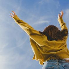 a woman with her arms up in the air while wearing jeans and a yellow sweater