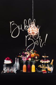 a table topped with cakes and drinks next to a chandelier that says bubbly bar