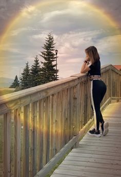 a woman is standing on a wooden bridge with a rainbow in the sky behind her