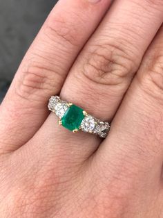 Elegantly displayed is a gorgeous three stone AAA emerald and diamond ring with accents. This romantic anniversary/engagement piece is captivating from every angle. The center features a Colombian emerald with Muzo green color and excellent eye clarity. This gem has a rich, dark green color that captivates at first glance. On either side, two brilliant round diamonds are flanked on either side of the emerald, totaling 0.46-carats. More round diamonds highlight the infinity styled shank and are t Elegant Three Stone Emerald Ring In White Gold, Elegant White Gold Emerald Three-stone Ring, Elegant White Gold Emerald Ring With Three Stones, Elegant White Gold Emerald Three Stone Ring, Luxury Three Stone Diamond Emerald Ring, Three-stone Emerald Diamond Ring As Gift, Three Stone Emerald And Diamond Ring For Gift, Three-stone Emerald Ring With Diamonds As A Gift, White Gold Emerald Diamond Ring With Three Stones