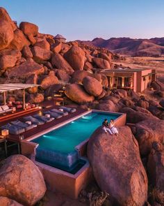 two people sitting on the edge of a swimming pool surrounded by rocks and boulders at dusk