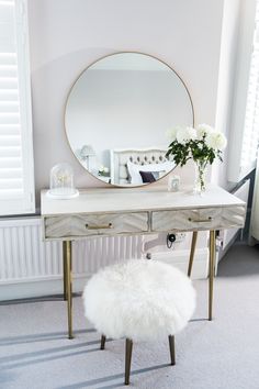 a white table with a mirror, stool and flowers on it in front of a window