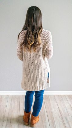 a woman standing in front of a white wall wearing a sweater and jeans with brown boots