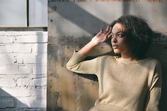 a woman in a tan sweater leaning against a brick wall with her hand on her head