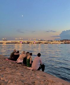 four people sitting on the edge of a river watching the sun go down in the distance