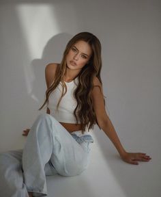 a beautiful young woman sitting on top of a white floor next to a wall and posing for the camera