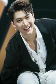 the young man is smiling and wearing a black suit with white shirt, jeans and gold necklace